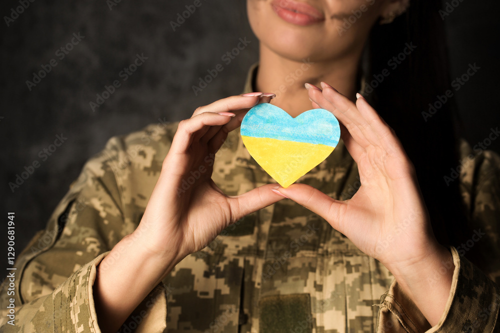 Military woman in military uniform with camouflage makeup on her face holds a heart-shaped Ukrainian flag in her hands. Close-up