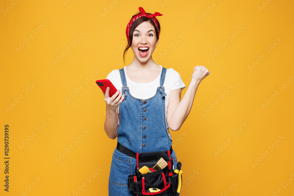 Young employee laborer handyman woman wear overalls belt hold use mobile cell phone do winner gesture isolated on plain yellow background Instruments for renovation apartment room Repair home concept Young employee laborer handyman woman wear overalls belt hold use mobile cell phone do winner gesture isolated on plain yellow background Instruments for renovation apartment room Repair home concept