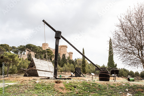 Una réplica de una máquina de asedio medieval hecha de madera, con un diseño imponente y robusto, se exhibe en un entorno natural cercano a un castillo.  photo