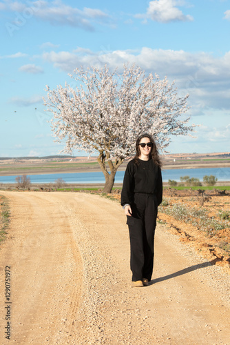 Una mujer vestida de negro con gafas de sol posa en un camino de tierra junto a un árbol en flor, con un hermoso paisaje natural de fondo que incluye un lago y colinas bajo un cielo azul con nubes. photo