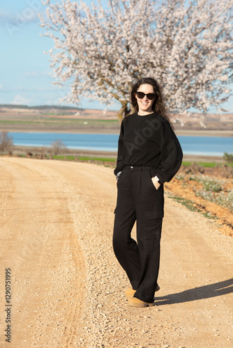 Una mujer vestida de negro con gafas de sol posa en un camino de tierra junto a un árbol en flor, con un hermoso paisaje natural de fondo que incluye un lago y colinas bajo un cielo azul con nubes. photo