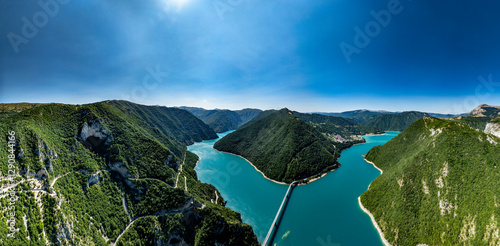 panorama Drone view of a bridge crossing the blue waters of Piva Lake between mountains in Montenegro, near a winding mountain road