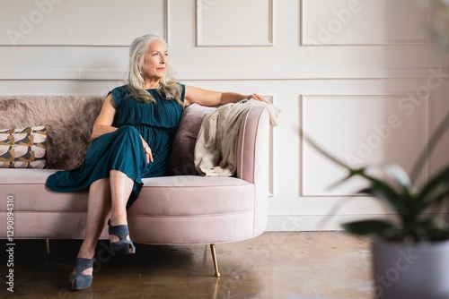 Elegant older woman wearing dress, sits on couch. The older woman looks thoughtful. Elegace old woman in luxury living room, calm old woman sitting thoughful, elderly woman in luxury vibes photo