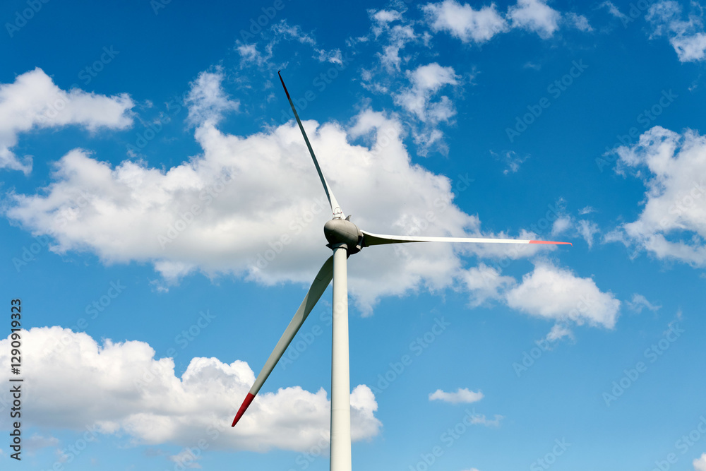 Modern wind turbine with red tipped blades stands tall against vivid blue sky filled with scattered white clouds, reflecting blend of technology and natural harmony Modern wind turbine with red tipped blades stands tall against vivid blue sky filled with scattered white clouds, reflecting blend of technology and natural harmony
