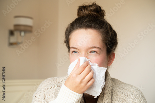 Sick young woman with a runny nose using a handkerchief, sitting in bed being unwell in the bedroom at home. Portrait of girl with fever, seasonal flu, cold, illness, viral infection. Home treatment.