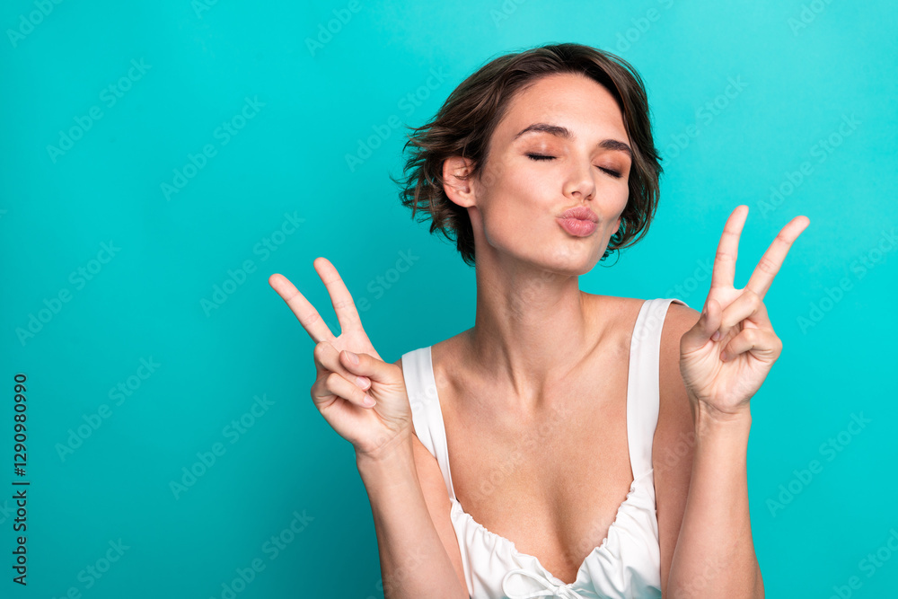 Fototapeta premium Charming young woman in casual top making a playful peace sign gesture against a vibrant teal background, exuding charm and positivity.
