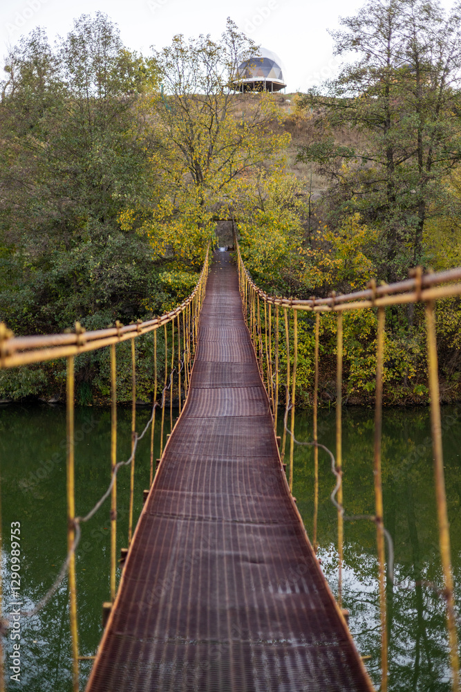 A narrow suspension bridge stretches through a lush autumn forest, surrounded by yellow leaves. The rusty metal path, covered with fallen leaves, leads into the distance, creating a mysterious.