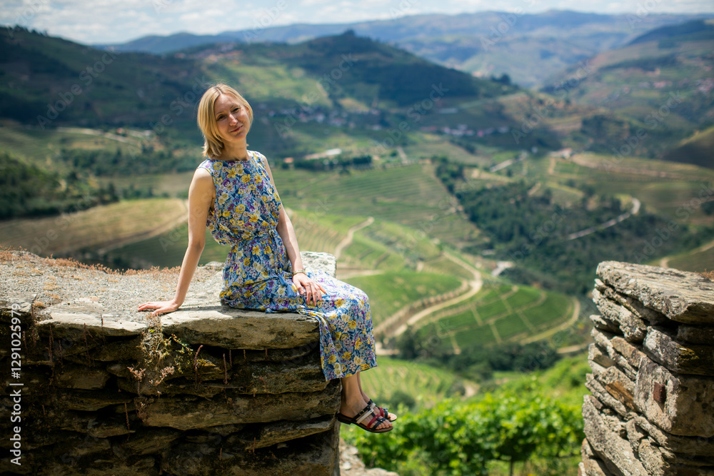 A woman in a light dress sits on a stone wall of a vineyard in the Douro Valley, Portugal.