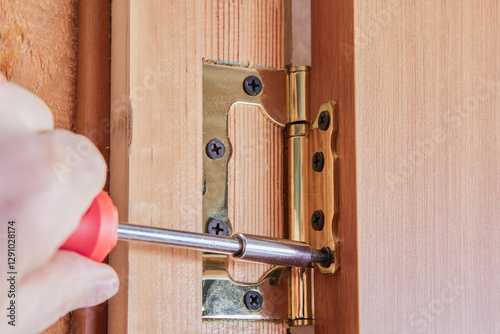 Screwdriver inserts screw into hinge leaf secured on door frame, demonstrating door installation technique with visible wood grain and aligned metal hardware.