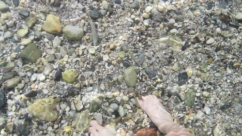 Bare female feet stand on a pebble-covered seabed as a school of fish swims nearby through crystal-clear water. 4K video 43 sec. Sunlight creates rippling patterns and rainbow reflections on the water