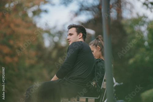 A young man is seated on a park bench outdoors, appearing thoughtful, with green foliage and a blurred background ambiance, capturing a serene autumn setting. photo