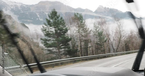 Navigating treacherous mountain road, windshield wipers clearing rain and snow while low clouds obscure dramatic alpine landscape during challenging winter driving conditions