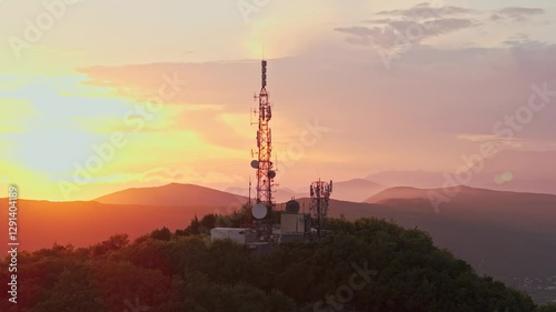 Aerial drone footage of a telecommunications tower on a green hill at sunset. Antennas and satellite dishes highlight modern connectivity, with mountains in the background. Perfect for themes of 5G, n