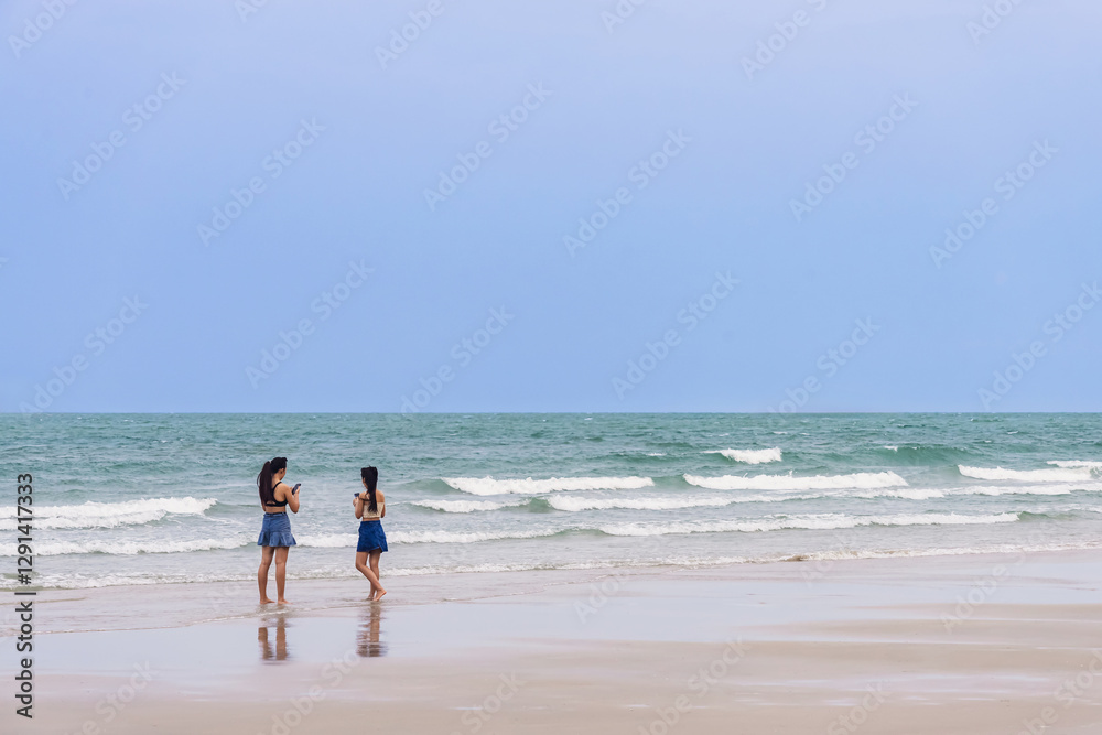 Back view of happy carefree women using mobile phone at beach. Woman friends using smartphone at seaside. Beautiful lovely two women spending time at beach. Concept of travel, tourism and vacation. Back view of happy carefree women using mobile phone at beach. Woman friends using smartphone at seaside. Beautiful lovely two women spending time at beach. Concept of travel, tourism and vacation.