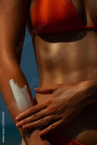 Female applying sunscreen on arm in a red bikini at the beach. Skin care, sun protection