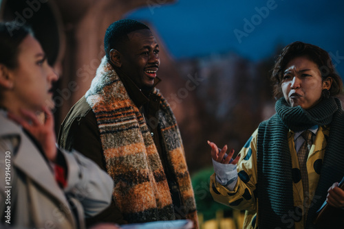 Casual conversation among diverse individuals outdoors in an urban setting, dressed warmly suggesting cooler weather. The evening environment enhances the scene's relaxed and friendly tone. photo