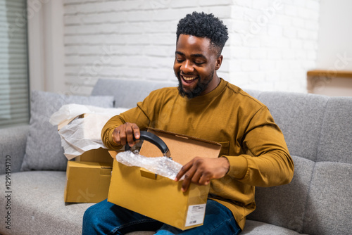 Young black man sitting comfortably in living room and unboxing package with joy after receiving online shopping delivery. Representation of customer satisfaction with home shopping.