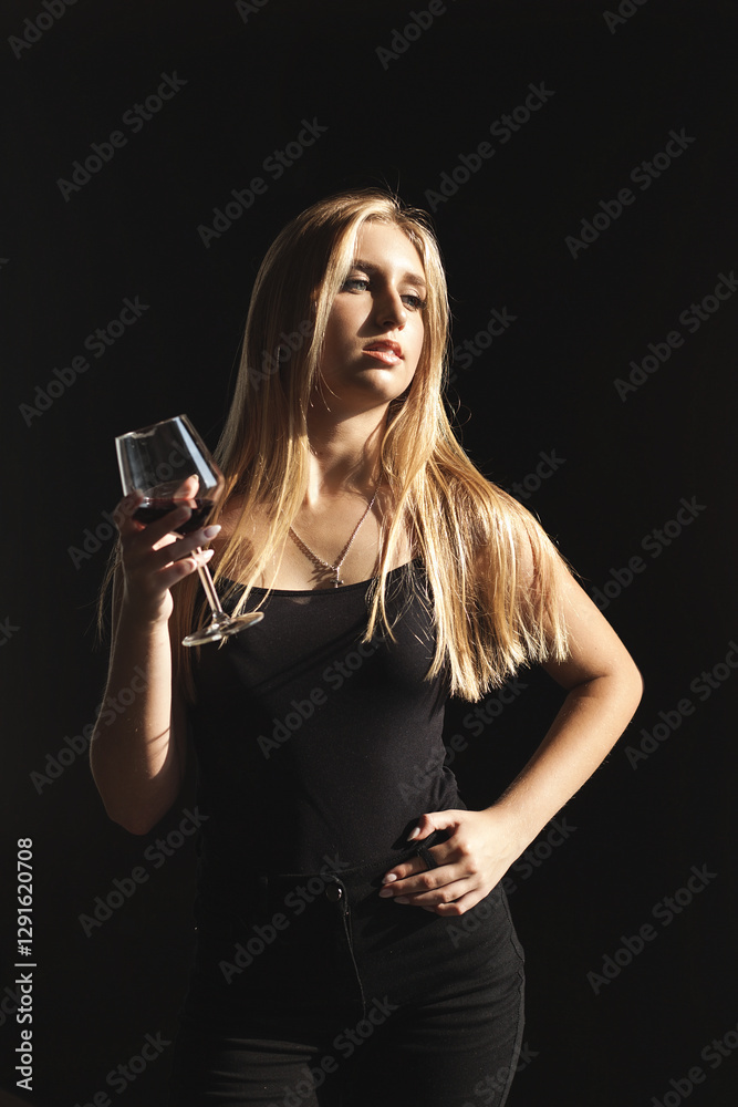 a woman with long blond hair stands against a dark background. She is wearing a black top and black pants. She holds a glass of red wine in one hand and places her other hand on her hip. a woman with long blond hair stands against a dark background. She is wearing a black top and black pants. She holds a glass of red wine in one hand and places her other hand on her hip.