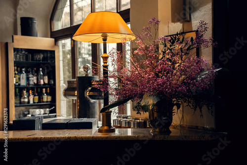 A warmly lit bar interior with an elegant table lamp, a lush bouquet of pink flowers, and a stylish bar counter. Perfect for hospitality, luxury, and lifestyle imagery.