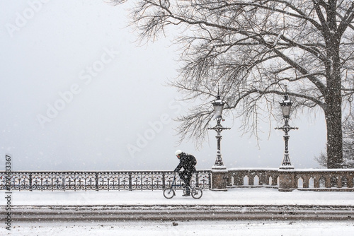 Ein Fahrrad fährt bei Winterbedingungen über eine Brücke mit zwei Laternen in Hamburg, an der der Alster, mit Schnee auf der Straße und Schneeflocken in der Luft photo