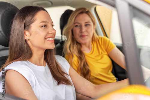 Obraz na płótnie Two women sit in a yellow car, sharing a joyful moment as the driver smiles confidently while her friend looks on with admiration, symbolizing friendship and adventure. w salonie