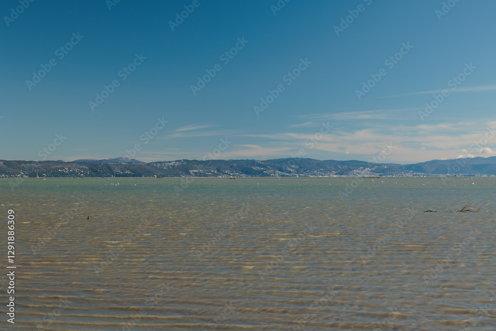 vista che si estende sull'area marina del Golfo di Trieste, nel mare Adriatico, guardando verso le colline e le montagne distanti tra Italia e Slovenia, di giorno, con cielo sereno, in inverno