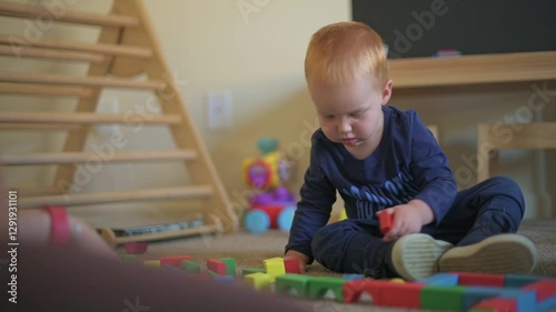 Focused Toddler Picking Up Colorful Block in Playroom young toddler sits on floor, carefully selecting colorful block while playing, developing motor skills and creativity in cozy indoor playroom.