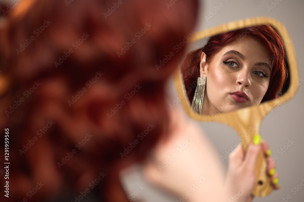 Reflection of woman with vibrant red hair in hand mirror, elegant earrings, stylish makeup, beauty and self-expression concept