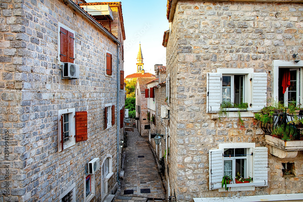 A charming stone alley in the ancient European town of Budva at sunset with a church spire in the distance.
