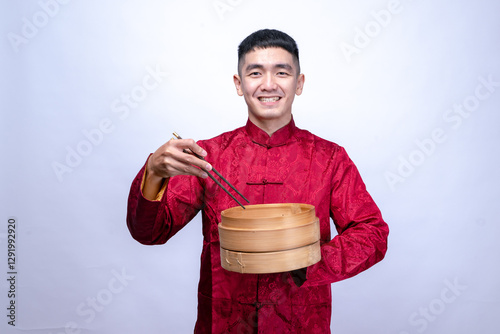 An Asian man in a red traditional Chinese Tang suit smiles while holding a bamboo steamer and using chopsticks, symbolizing Chinese cuisine, isolated against a plain white background photo