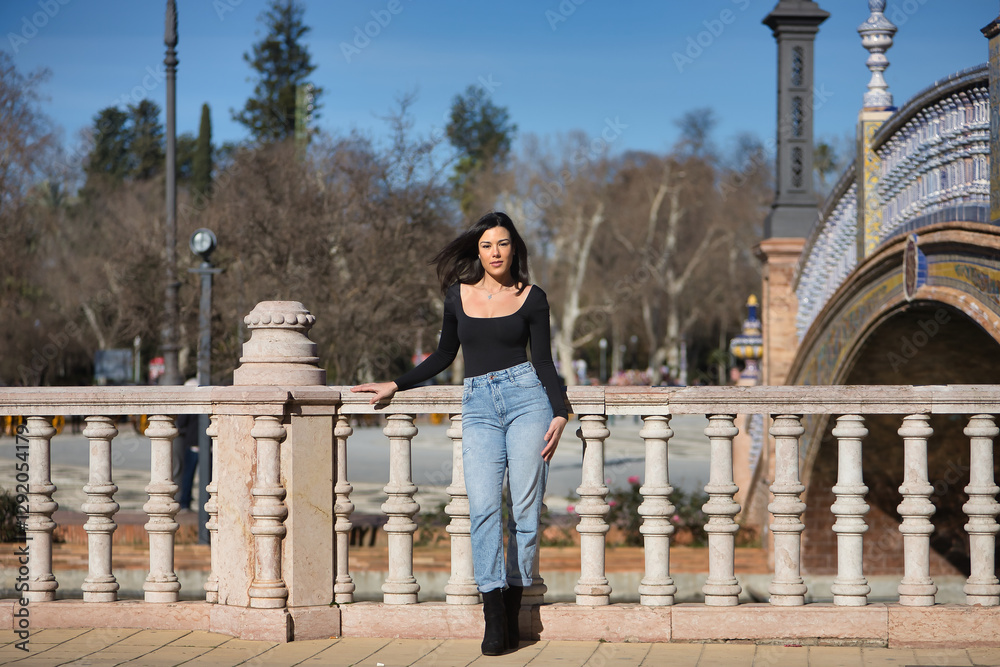 Fototapeta premium Young, beautiful, brunette woman, wearing black long sleeve t-shirt and jeans, with her hair in the wind, posing leaning on a railing in the square of Spain in Seville. Concept beauty, femininity.