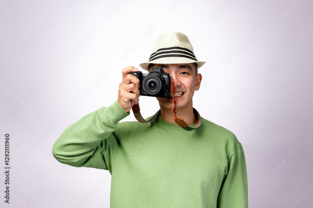 An Asian man wearing a fedora hat and a green sweater is holding a camera up to his eye, smiling confidently as if taking a picture. The white background enhances the focus on him