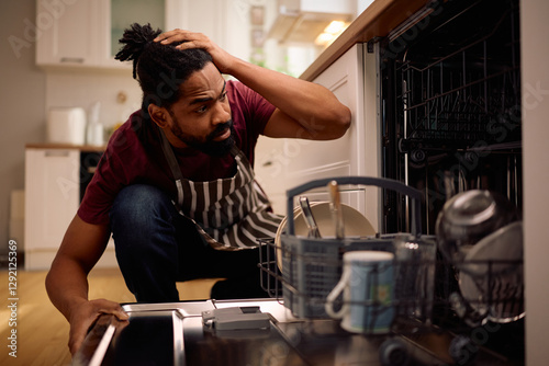 African American man feeling upset about dishwasher malfunctioning in kitchen.