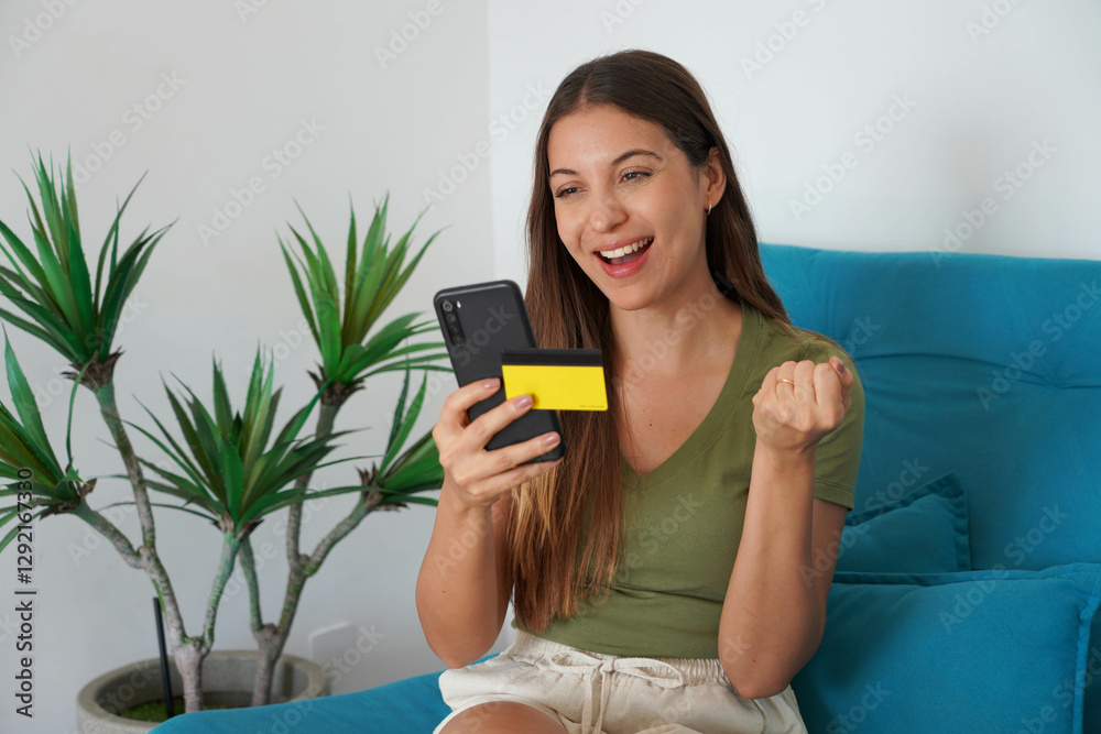 Happy young Brazilian woman holding smartphone and credit card celebrates with a clenched fist sitting on couch at home Happy young Brazilian woman holding smartphone and credit card celebrates with a clenched fist sitting on couch at home