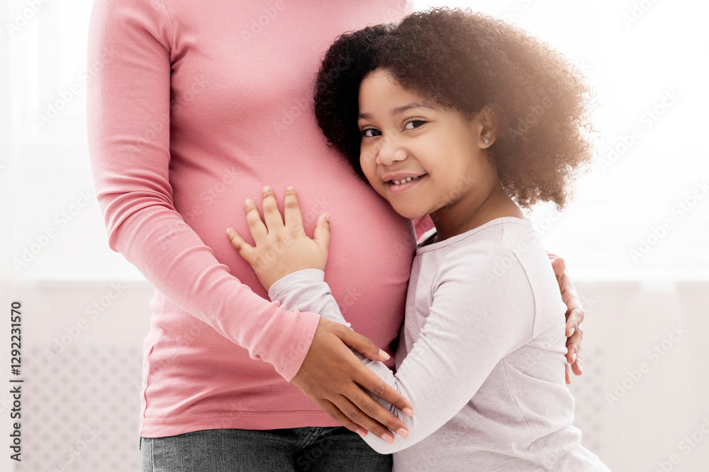 Fototapeta premium Close up of little black daughter hugging her pregnant mother's tummy, standing next to window at home