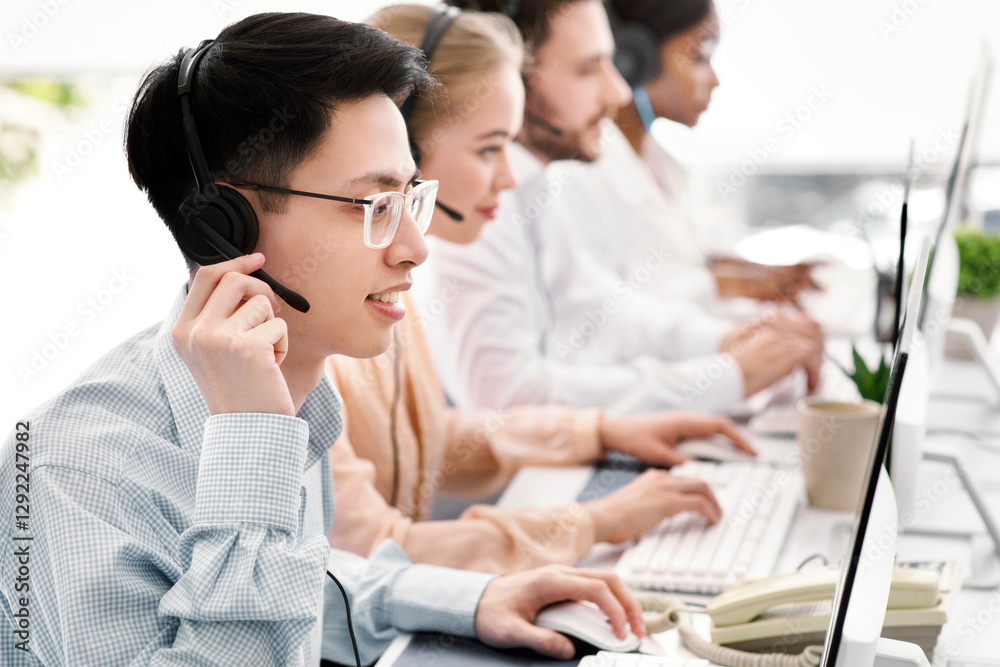 Asian helpdesk representative in headphones with his colleagues providing tech support services to customers, copy space. Team of call centre managers communicating with clients or selling online Asian helpdesk representative in headphones with his colleagues providing tech support services to customers, copy space. Team of call centre managers communicating with clients or selling online