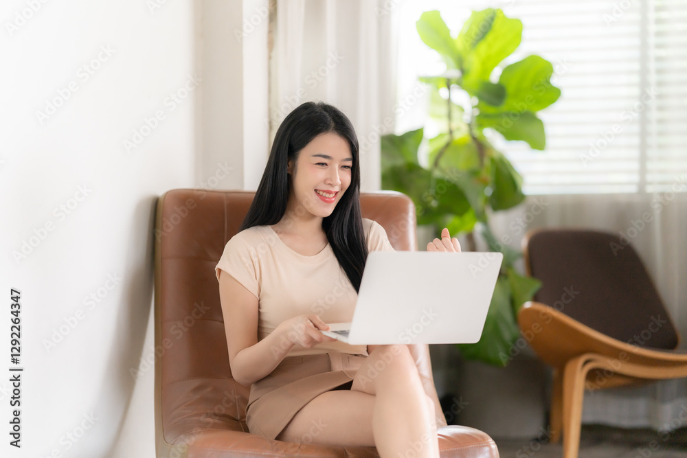 Woman Working Remotely: A young woman is seated indoors, using a laptop computer, in a casual setting with a happy expression. Woman Working Remotely: A young woman is seated indoors, using a laptop computer, in a casual setting with a happy expression.