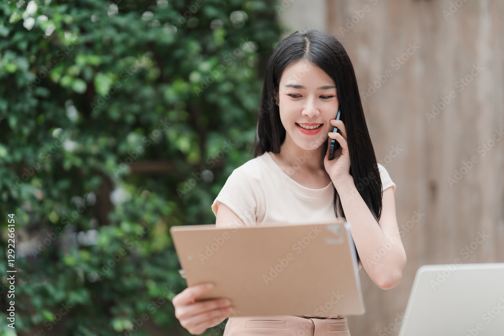 Business Woman on a Phone: A cheerful Asian businesswoman holds a phone to her ear while reviewing documents. She is near plants. Business Woman on a Phone: A cheerful Asian businesswoman holds a phone to her ear while reviewing documents. She is near plants.