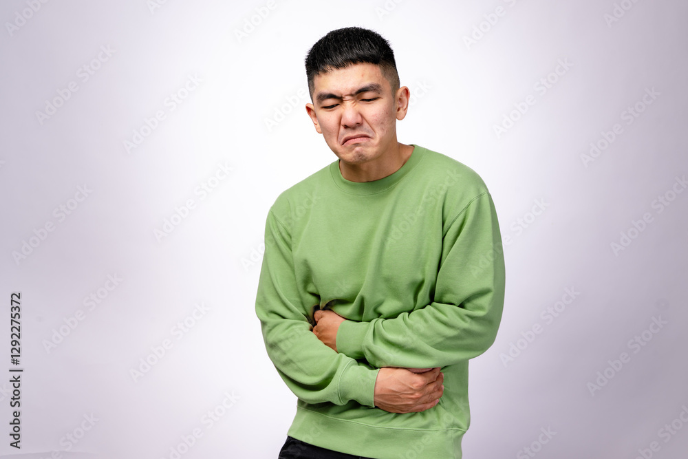 An Asian man in a green sweatshirt is grimacing in pain, holding his stomach and lower back. His facial expression suggests intense discomfort. The background is plain white