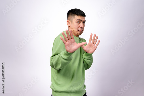 An Asian man in a green sweatshirt makes a hesitant expression while raising both hands in a defensive gesture. His eyebrows are raised, and his mouth is slightly pouted. The background is plain white
