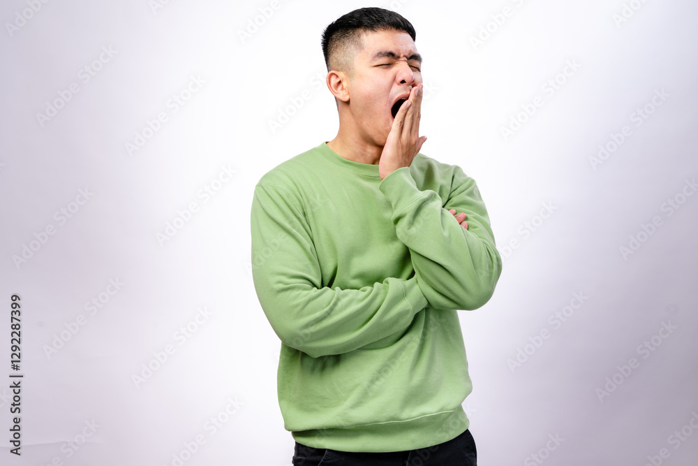 An Asian man wearing a green sweatshirt stands against a white background, yawning with his hand covering his mouth. He has his arms crossed, expressing tiredness or boredom An Asian man wearing a green sweatshirt stands against a white background, yawning with his hand covering his mouth. He has his arms crossed, expressing tiredness or boredom