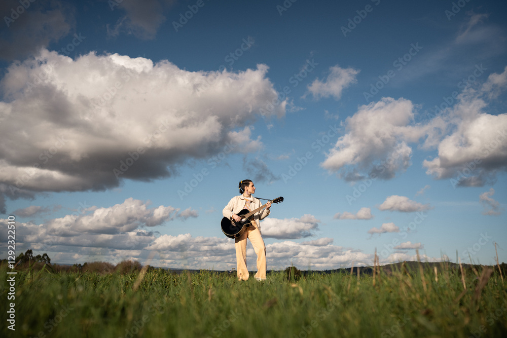 Obraz premium Woman playing acoustic guitar in grassy field beneath blue sky, white clouds surrounding her peaceful musical moment in sunlit outdoor setting