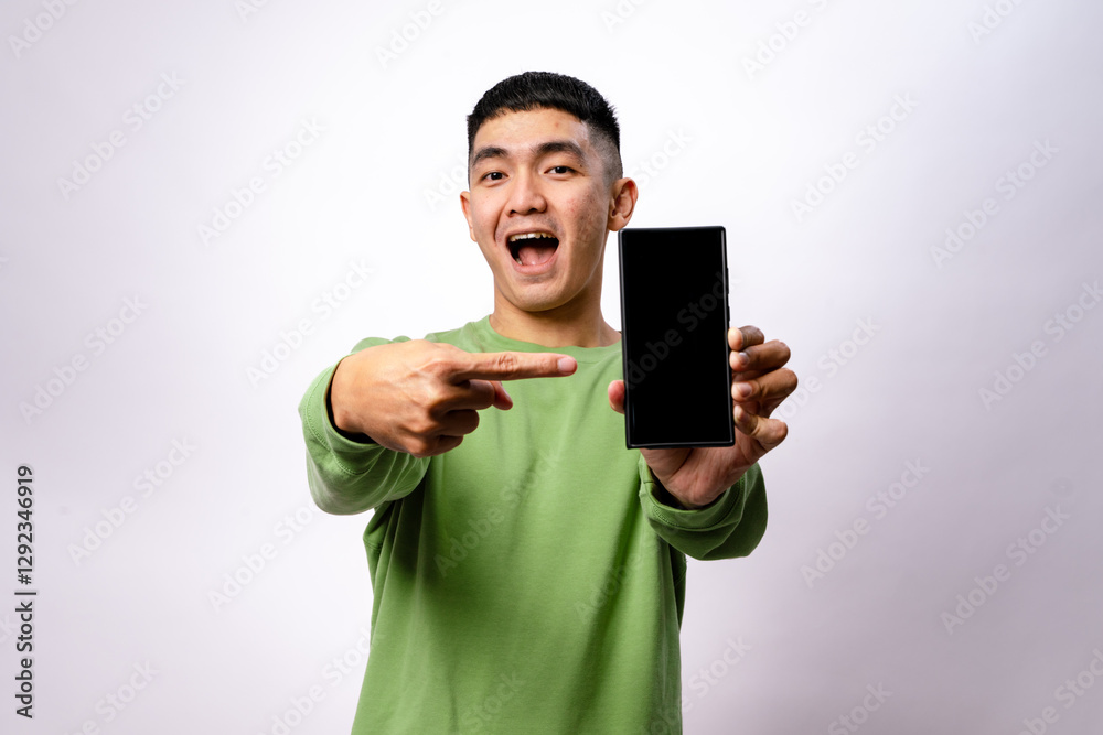 An Asian man in a green sweatshirt is smiling and pointing at a smartphone with a blank screen. He holds the phone in one hand while gesturing towards it with excitement. The background is white An Asian man in a green sweatshirt is smiling and pointing at a smartphone with a blank screen. He holds the phone in one hand while gesturing towards it with excitement. The background is white