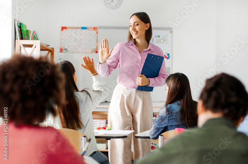 Friendly young European female teacher giving high five to pupil, standing near her desk and smiling. Education, teaching, learning at private school concept photo