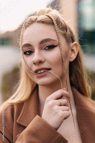 Young blonde woman with braided hair, wearing a stylish brown coat and a ring, playfully holding a strand of her hair between her teeth, exuding confidence and charm