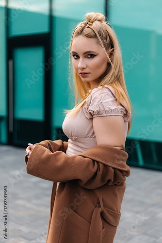Young blonde woman wearing a stylish brown coat and a beige top, showcasing a braided hairstyle while posing confidently in front of a striking turquoise modern building