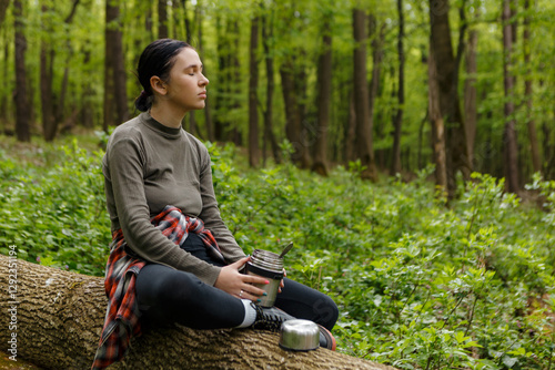 Young hiker woman is enjoying a peaceful moment in the forest during a hike break, sitting on a fallen tree trunk and eating from a thermos