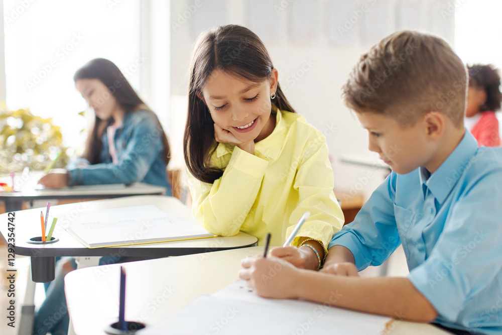 Positive schoolgirl sitting at table near boy classmate and helping with task, writing in his copybook, enjoying learning process in school Positive schoolgirl sitting at table near boy classmate and helping with task, writing in his copybook, enjoying learning process in school