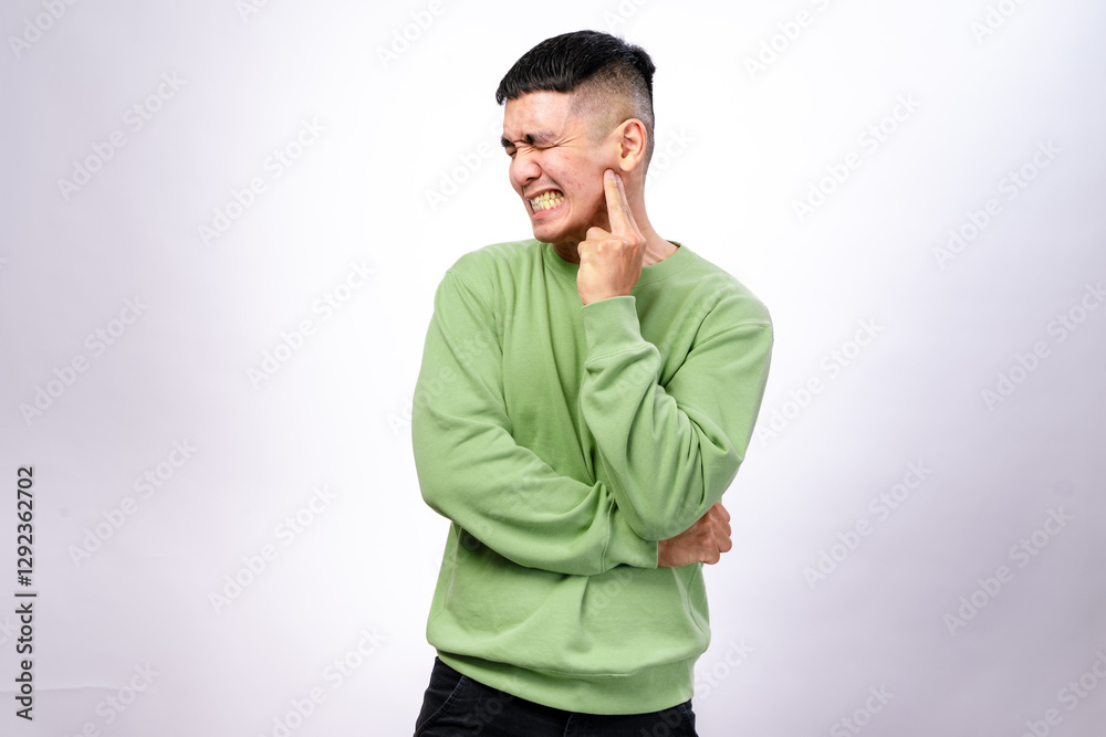 An Asian man in a green sweatshirt clenches his teeth and squints his eyes while pointing to his cheek, expressing pain. His facial expression suggests a toothache against a white background An Asian man in a green sweatshirt clenches his teeth and squints his eyes while pointing to his cheek, expressing pain. His facial expression suggests a toothache against a white background