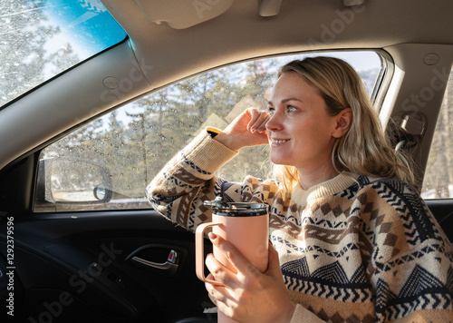 Young beautiful caucasian woman sitting in the car and drinking coffee from a pink mug, girl is smiling and dreaming looking forward through front windshield. Nice and sunny day for a trip